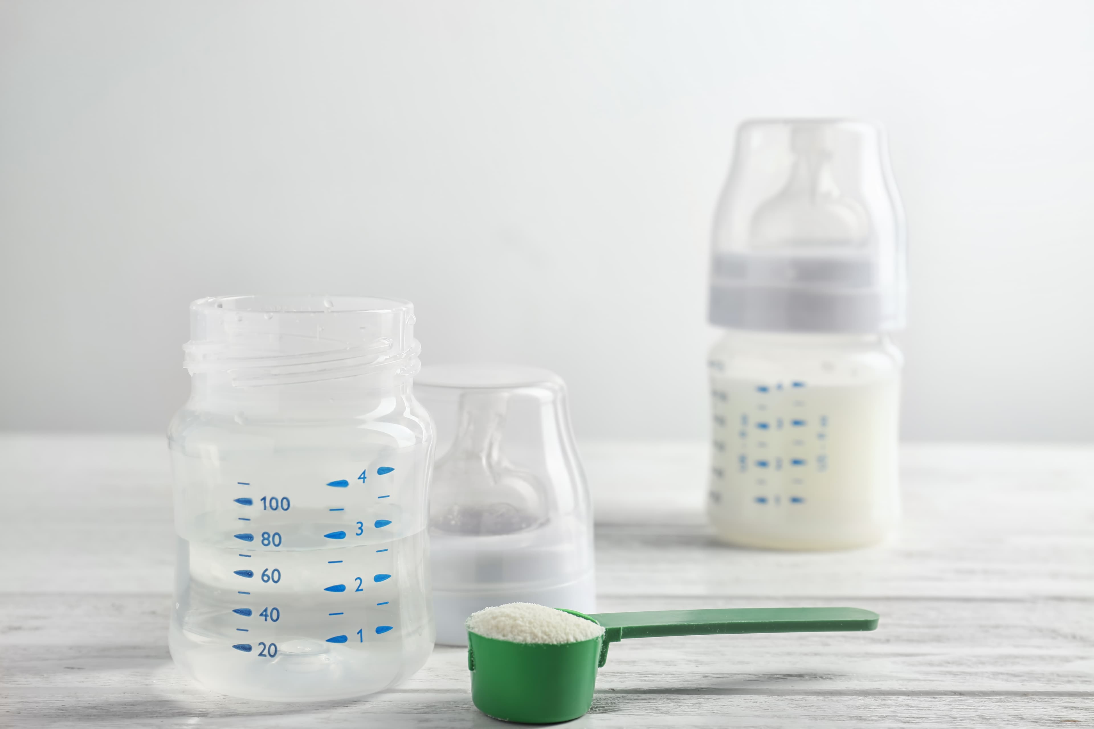 A green measuring scoop filled with powdered infant formula in front of an empty baby bottle with measurement markings, with a second bottle filled with prepared formula in the background.
