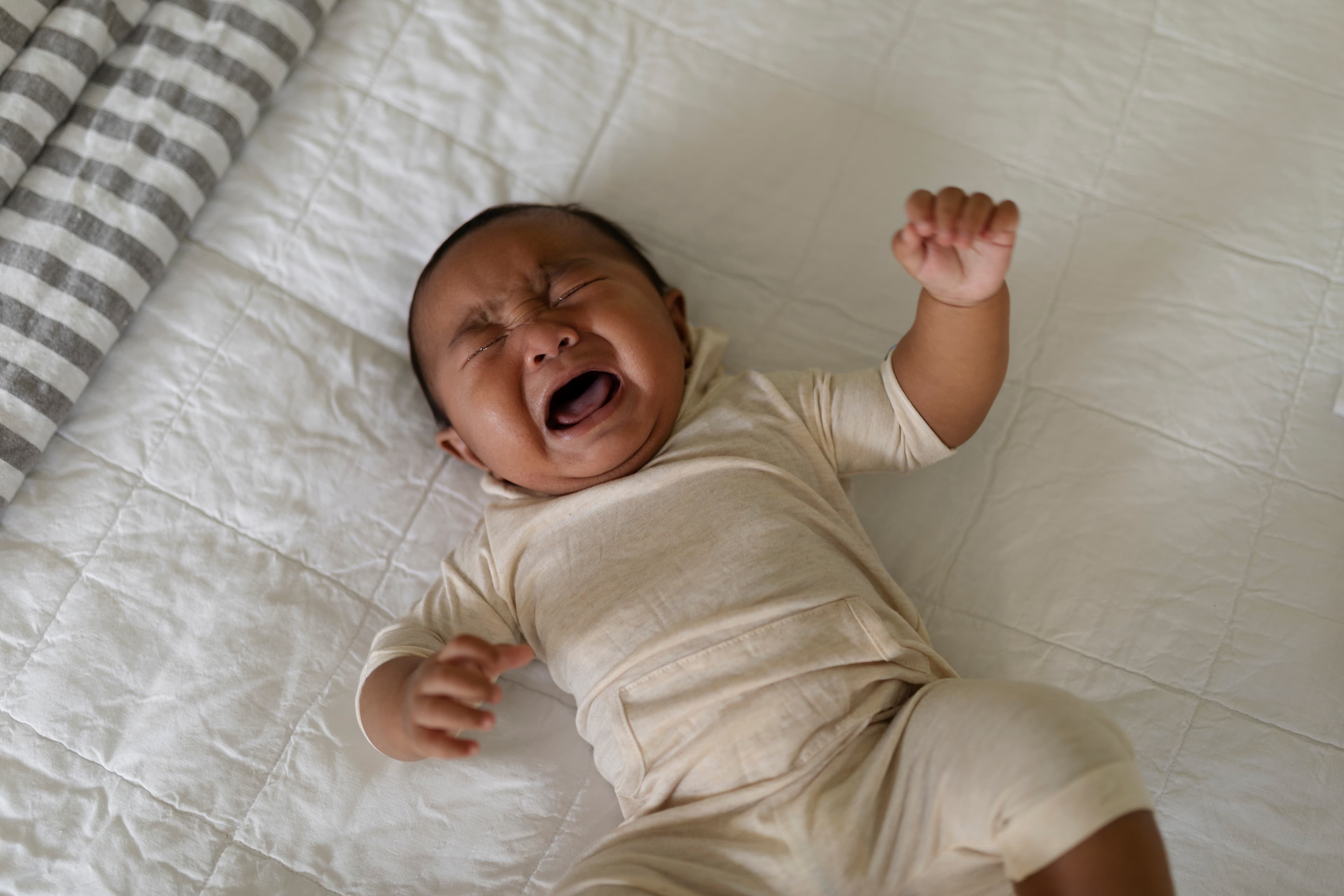 A baby in a cream-colored onesie lying on a white quilted surface, crying with eyes shut and fists raised.